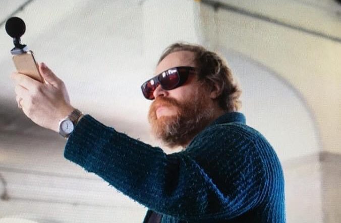 A white man with red hair and red wraparound glasses wearing a blue cardigan holding up a microphone in a white room. 