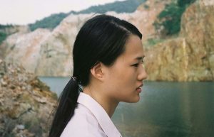 A Thai woman with a low ponytail wearing a white shirt standing in profile against a river landscape.