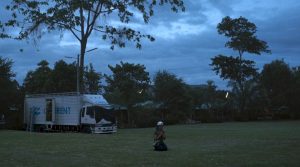 A Thai woman in a white backwards baseball cap kneeling in an outdoor field at dusk. Behind her is a white truck and tall trees.