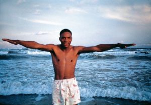 A black man in white swim trunks standing with arms outstretched on an ocean beach.