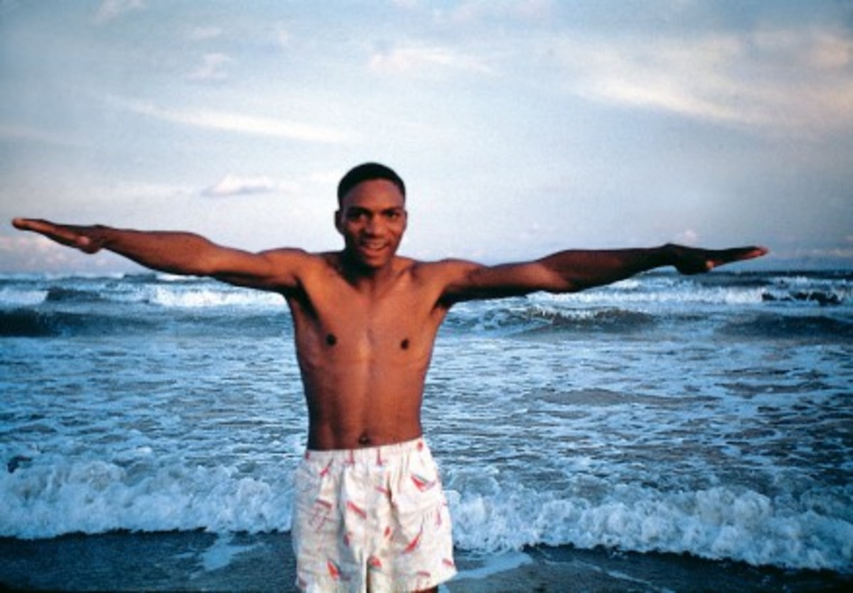 A black man with white swim trunks standing with arms outstretched on an ocean beach.
