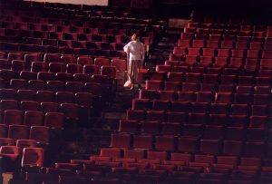 a woman with a red broom and dustpan sweeps an empty theater full of red seats.