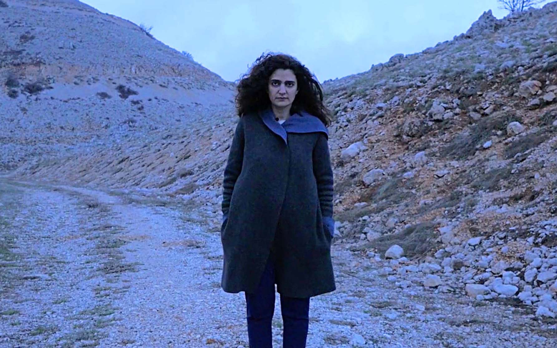 A women stands in a deserted landscape looking seriously at the camera.