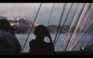 Two silhouettes look at land from behind the sail ropes of a ship. 