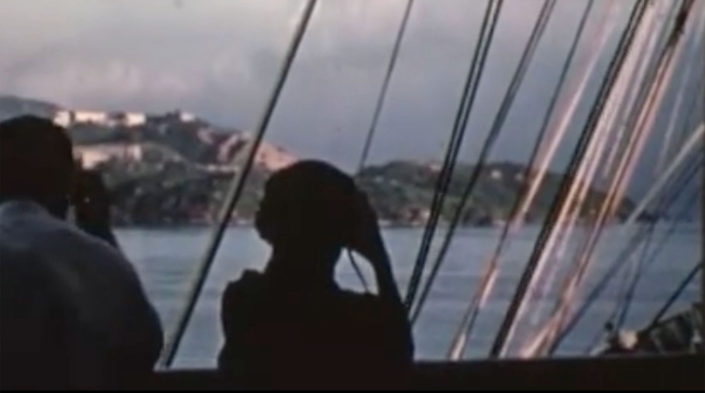 Two silhouettes look at land from behind the sail ropes of a ship. 