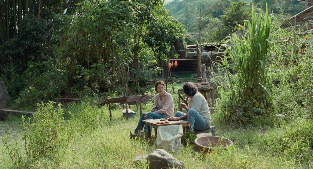 Two figures sitting at a table in a lush green landscape.