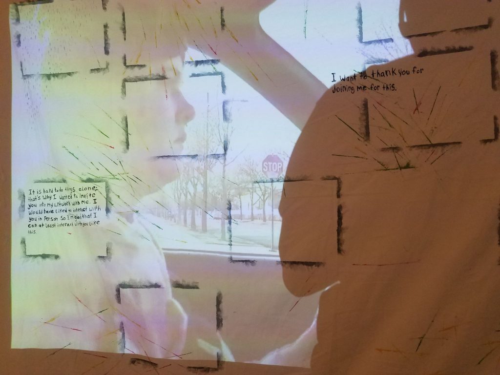 Video and Image Descriptions: The piece shows a video being projected onto a painted canvas. The canvas has partial outlines of rectangles that form a grid pattern with some spaces left without rectangles. There are also various spots on the canvas that show painted lines in various colors, gray and blue dots, and multi-colored clouds. There are two sections of writing as well. In a rectangle on the left there are the words 