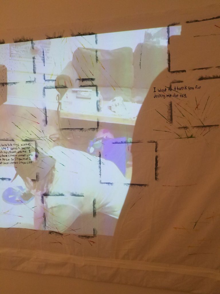 Video and Image Descriptions: The piece shows a video being projected onto a painted canvas. The canvas has partial outlines of rectangles that form a grid pattern with some spaces left without rectangles. There are also various spots on the canvas that show painted lines in various colors, gray and blue dots, and multi-colored clouds. There are two sections of writing as well. In a rectangle on the left there are the words 